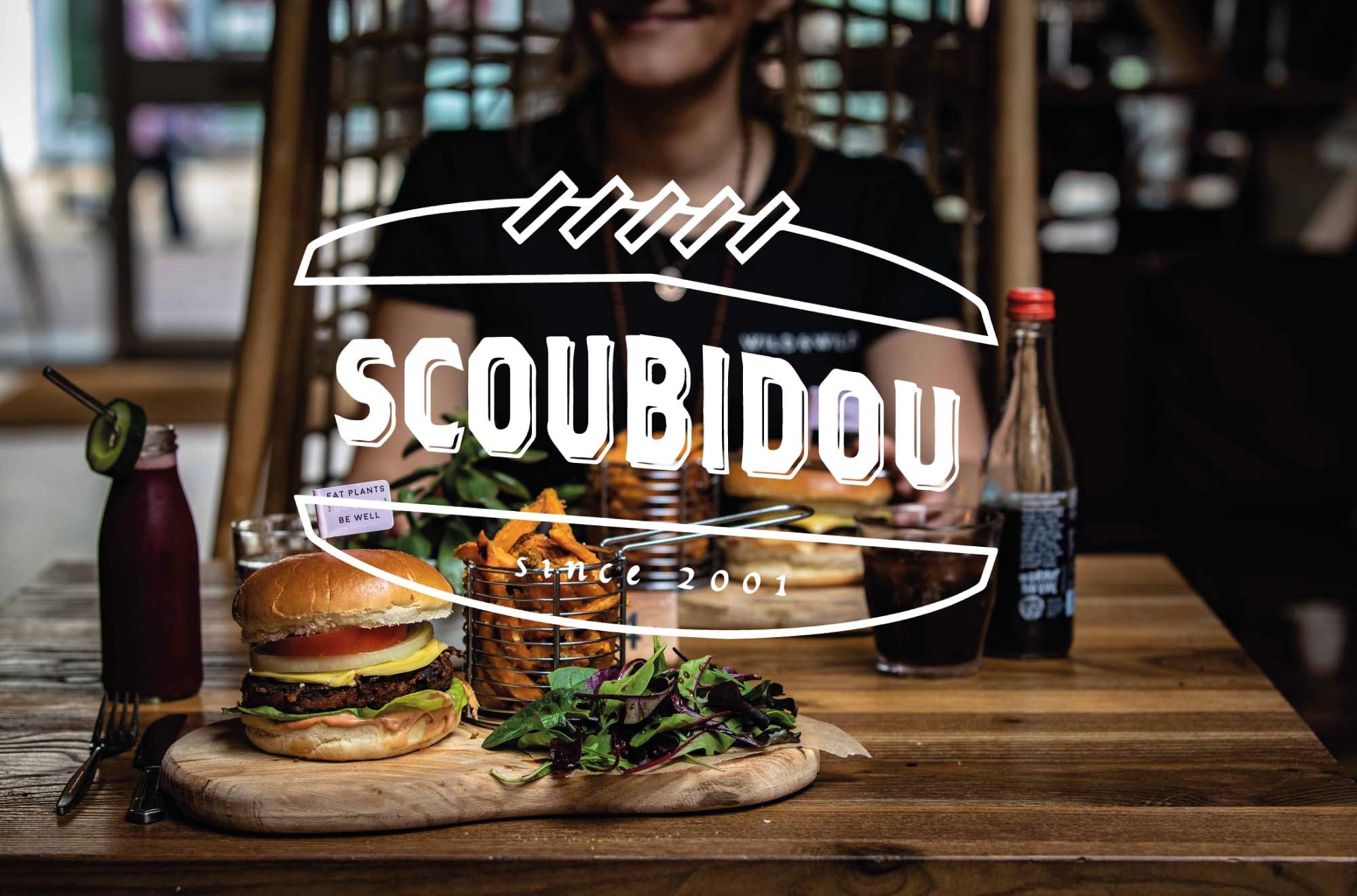 Logotype de Scoubidou réalisé par l'agence ÈS.B Studio en blanc sur fond photographique représentant une table de restaurant avec deux repas sur la table en couleur.